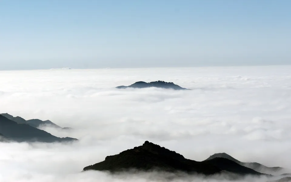 Silhouetted mountain peaks emerge from a thick blanket of white clouds under a clear blue sky.