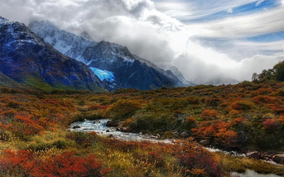 A winding river flows through a valley of autumn foliage towards snow-capped mountains with a bright blue glacier.
