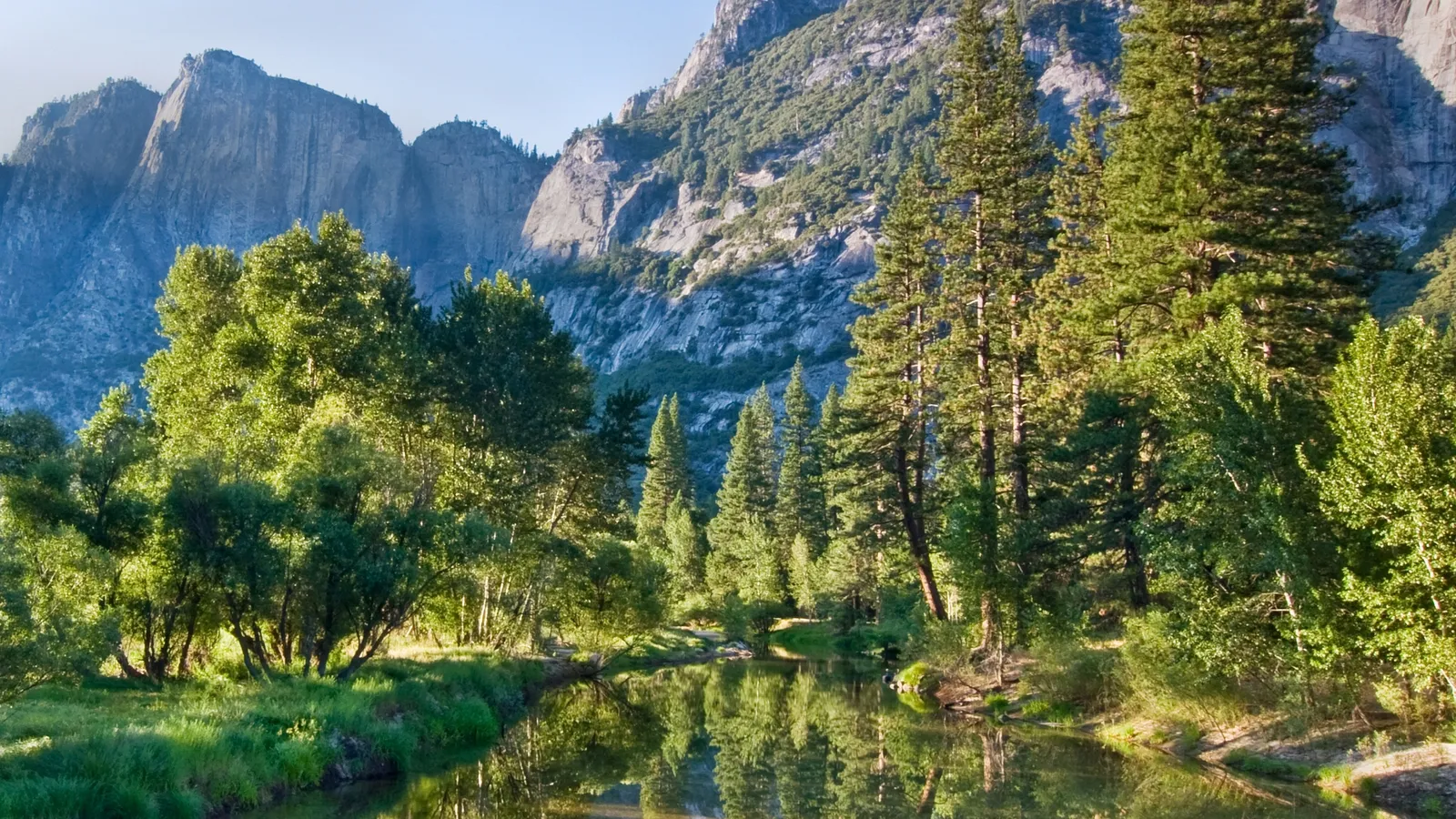A calm river reflects lush green trees and imposing granite mountains under a clear blue sky.