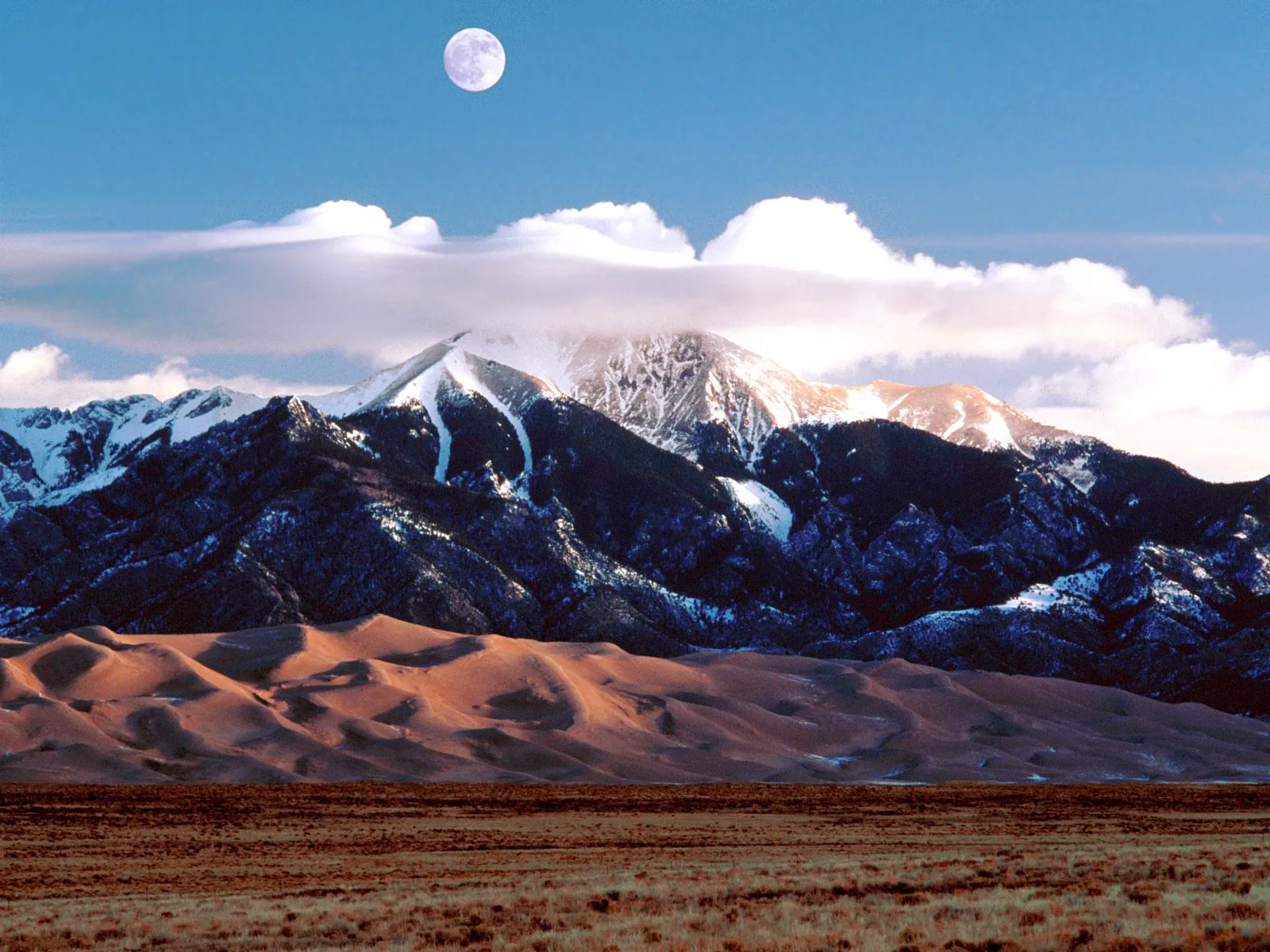 Snow-capped mountains rise above rolling sand dunes under a blue sky with a full moon.