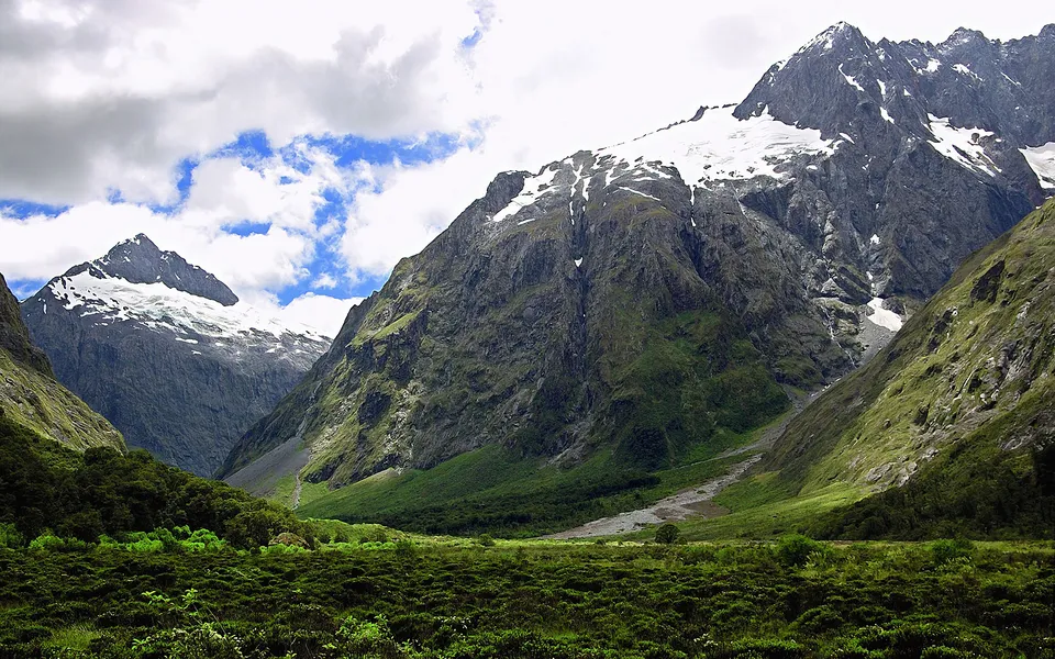 A wide valley with green vegetation leads to towering, rugged mountains capped with snow under a cloudy sky.