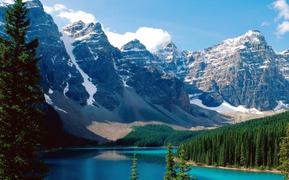 A stunning view of snow-capped mountains overlooking a vibrant turquoise lake surrounded by pine trees.