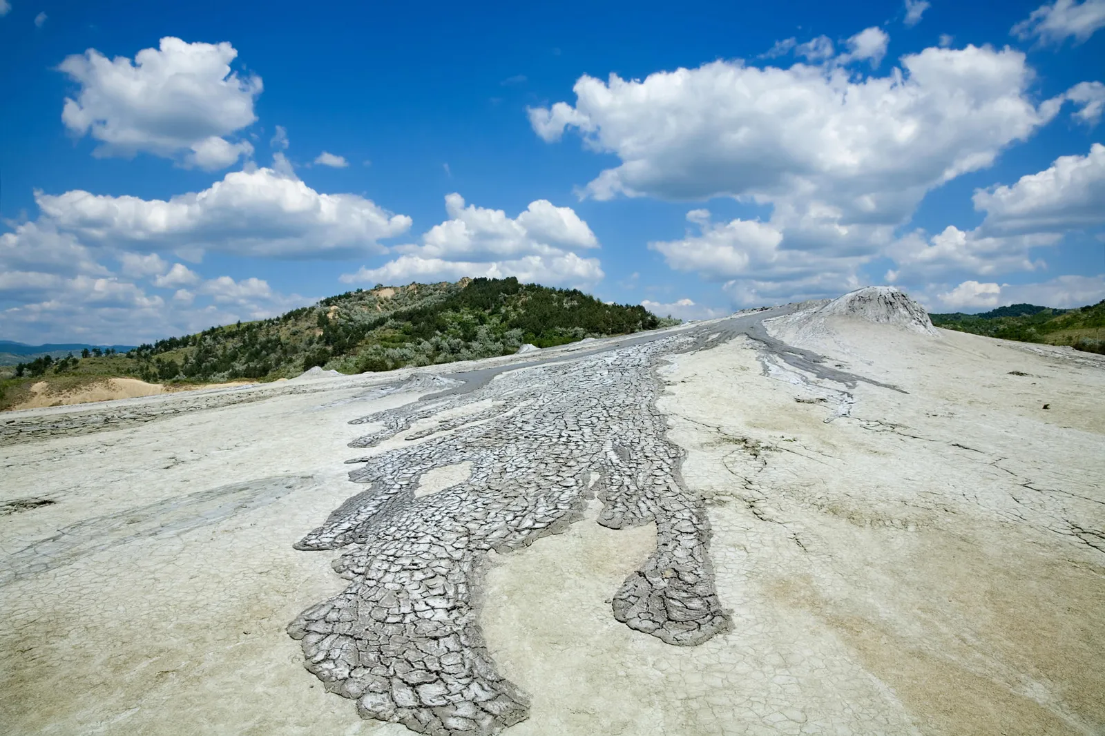 Mud volcanoes with cracked earth and flowing mud under a bright blue sky with white clouds.