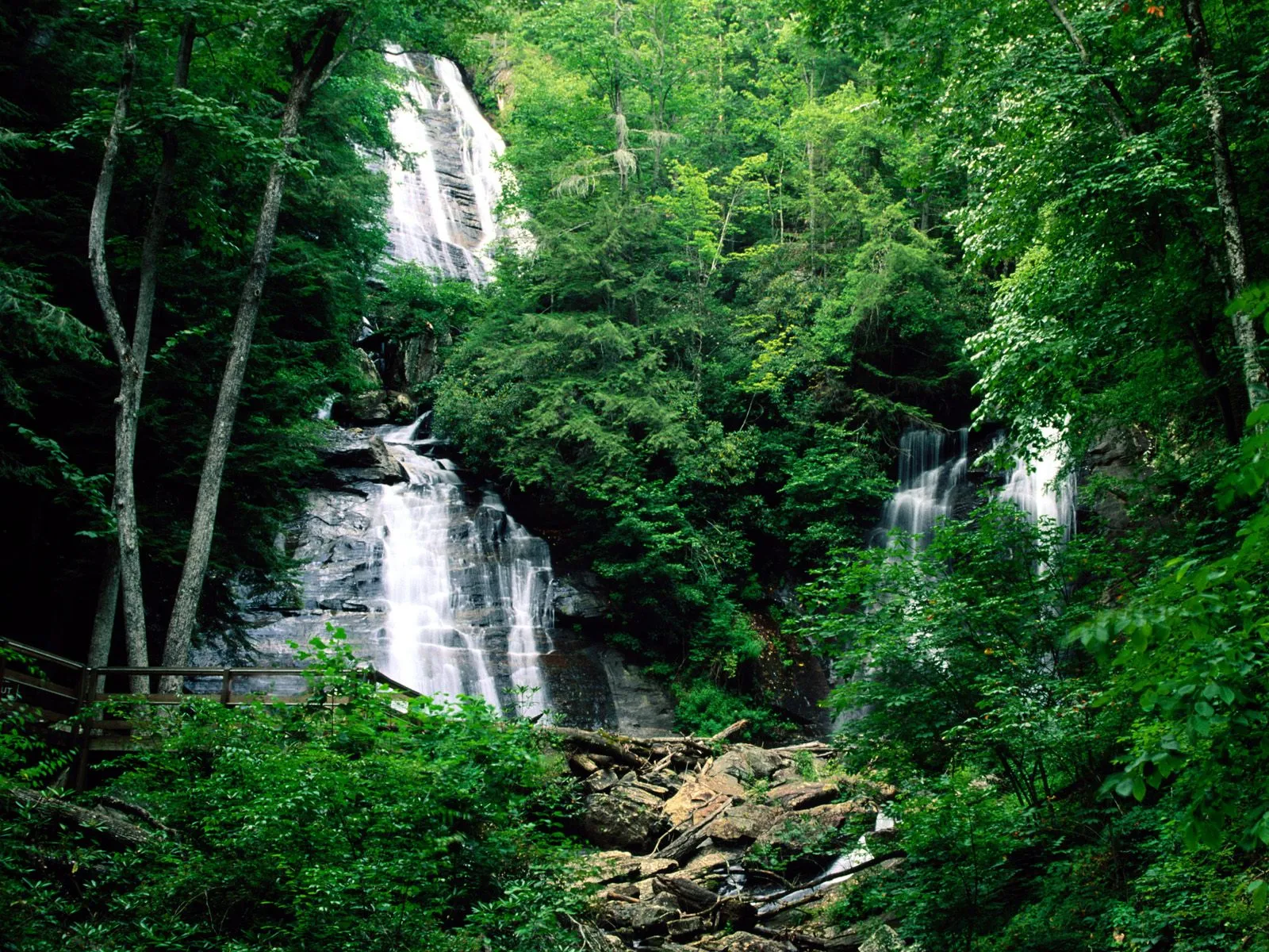 A multi-tiered waterfall flows down a rocky incline in a dense forest, with a wooden boardwalk visible on the left.