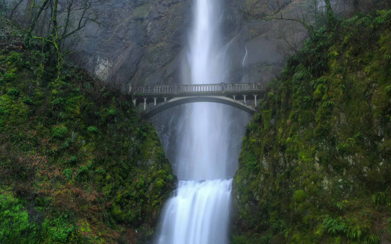 A tall, multi-tiered waterfall cascades down a rocky cliff face, with a historic arched bridge spanning across it.