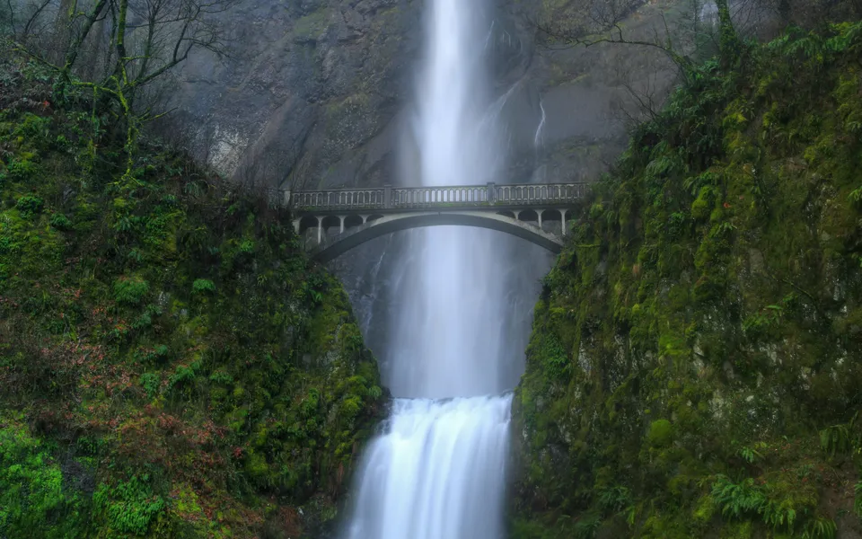 A tall, multi-tiered waterfall cascades down a rocky cliff face, with a historic arched bridge spanning across it.