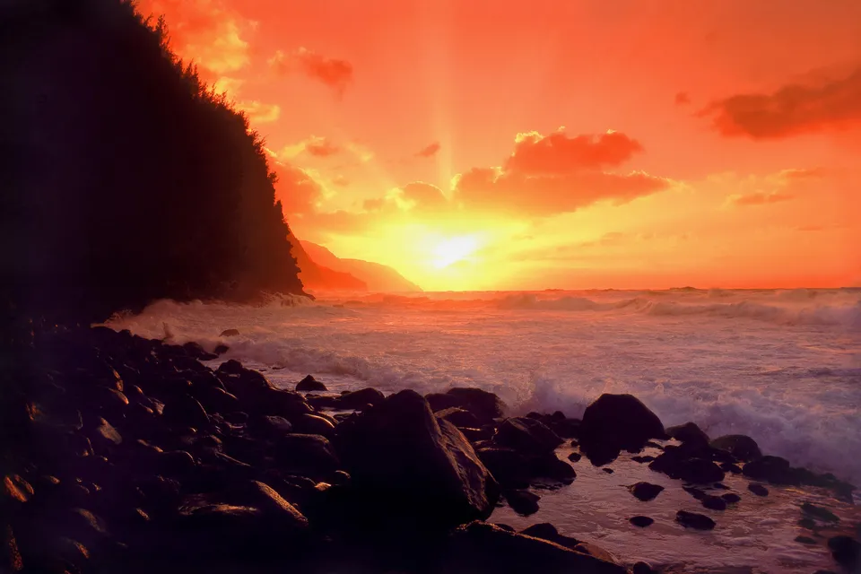 A vibrant sunset casts an orange glow over the ocean waves crashing against a rocky shore and a lush cliffside.