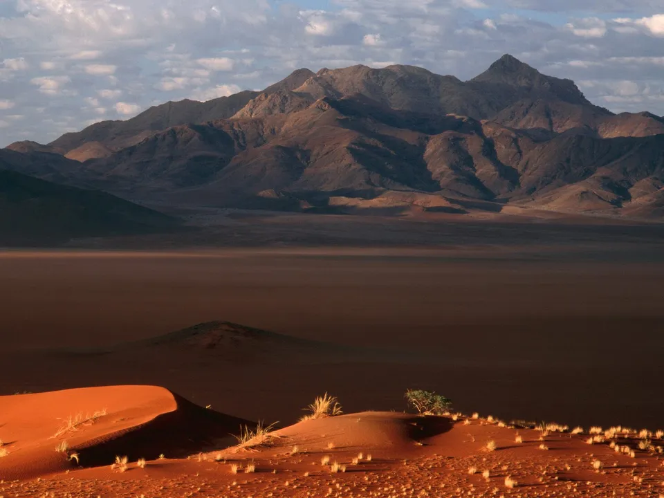 Vast desert landscape with red sand dunes in the foreground and rugged mountains in the background under a cloudy sky.