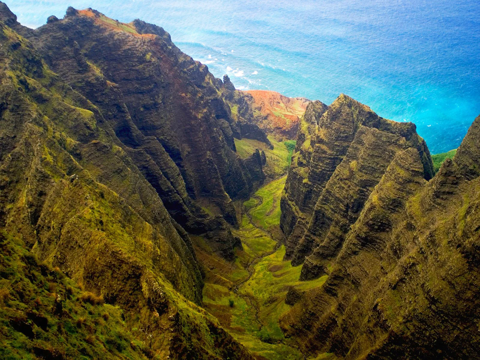 Aerial view of the rugged, green Napali Coast cliffs plunging into the bright blue ocean.