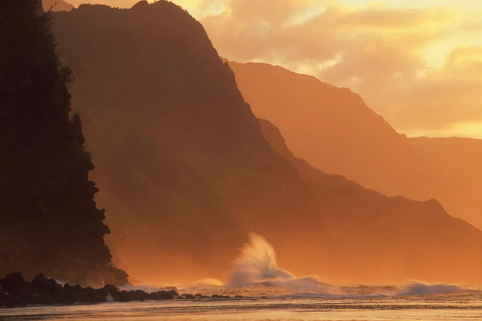 Waves crash against the rocky shore below towering, mist-shrouded mountains at sunset.