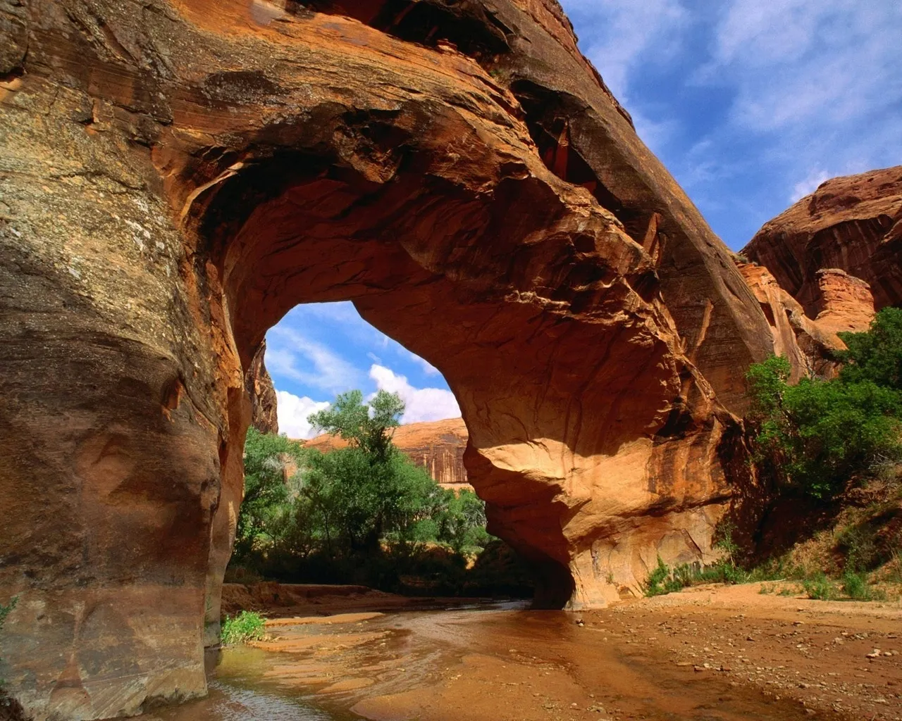 A natural sandstone arch frames a view of a desert stream and green trees under a blue sky.