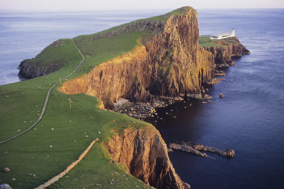 A winding path leads across green cliffs to a lighthouse on a rocky promontory overlooking the sea.