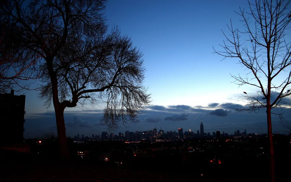 A silhouetted cityscape of New York City at dusk, with bare trees in the foreground against a deep blue sky.