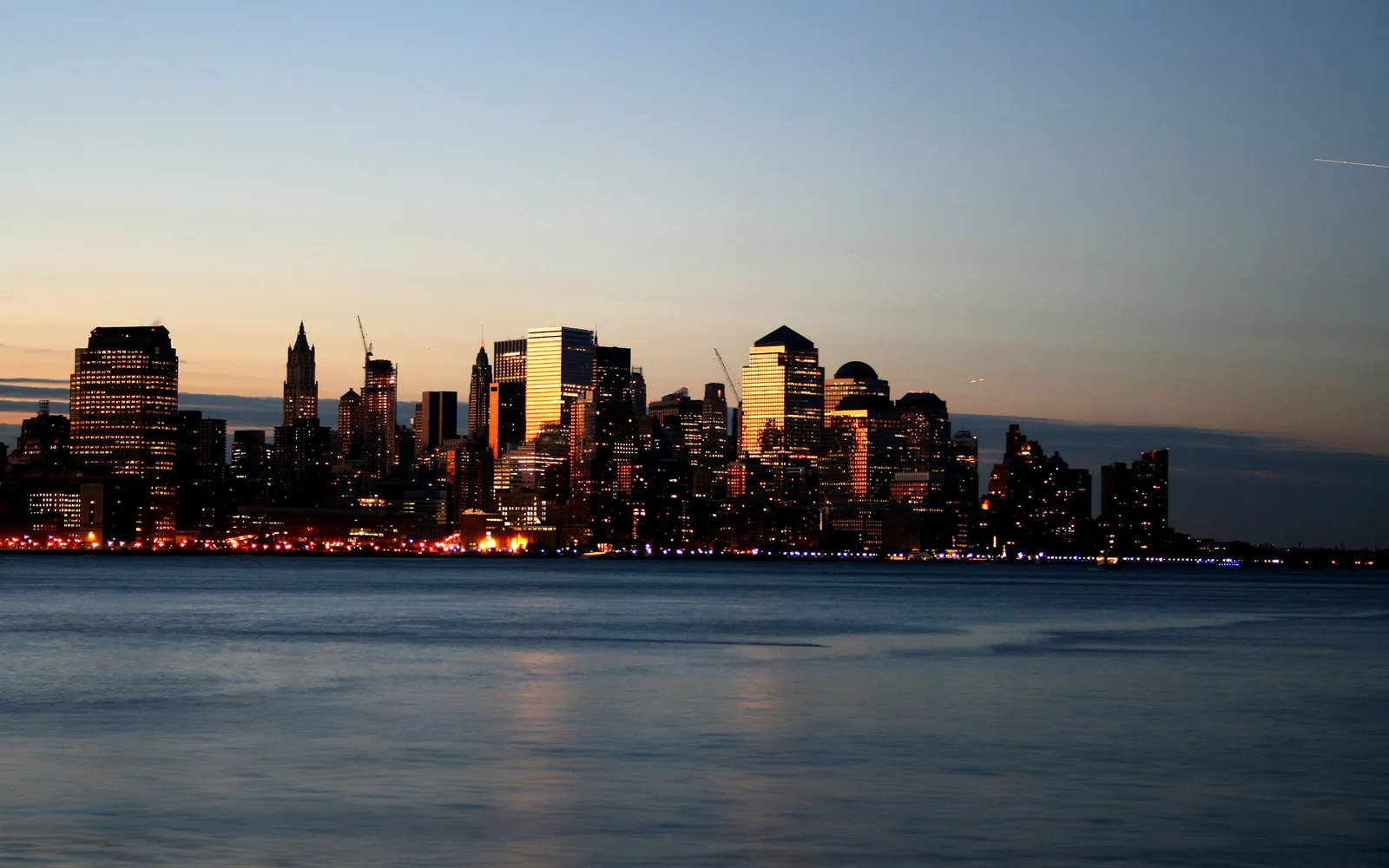 New York City skyline at dusk with buildings reflecting on calm water.