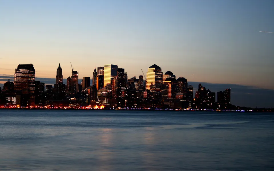 New York City skyline at dusk with buildings reflecting on calm water.