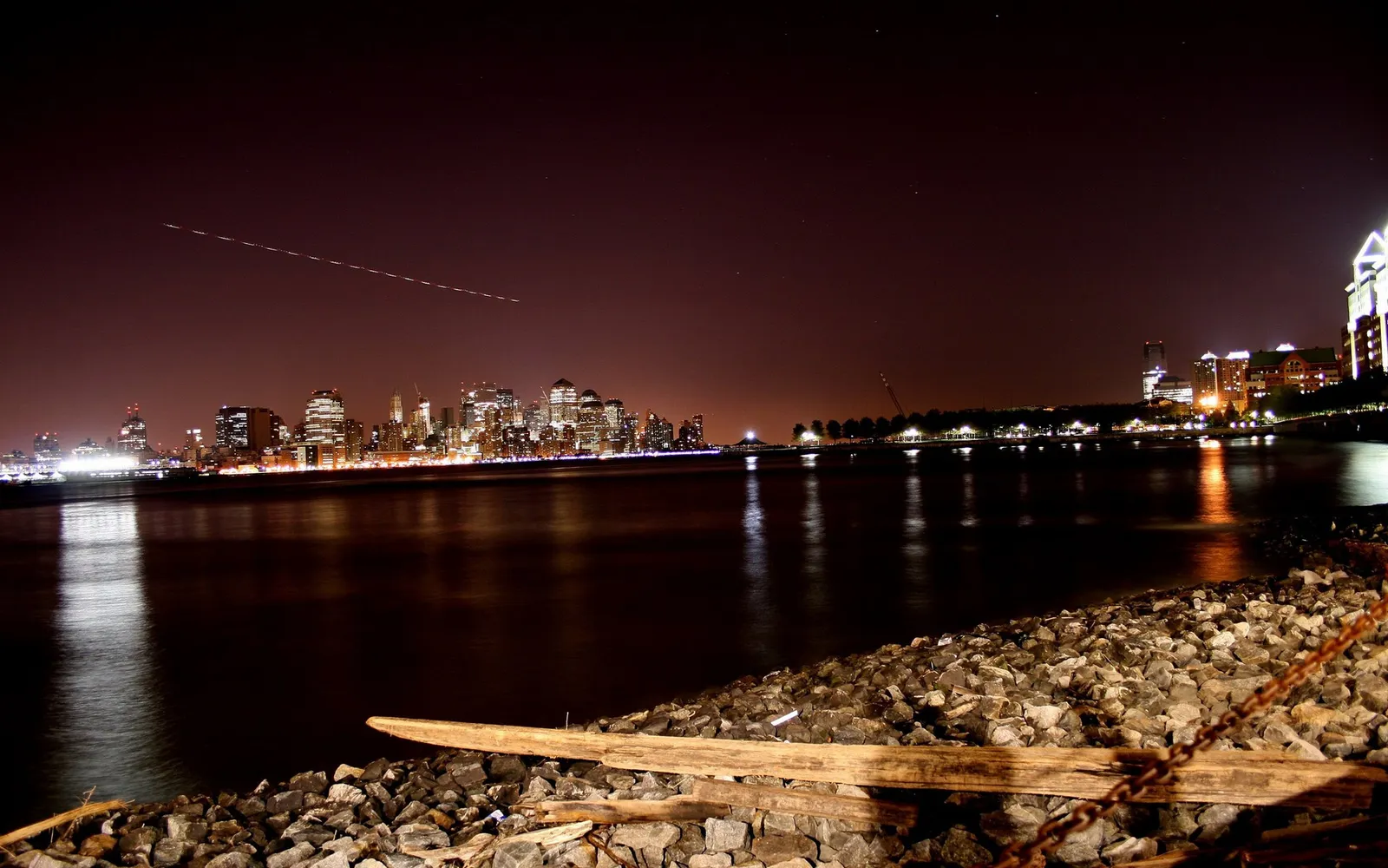 New York City skyline illuminated at night, reflected in dark water, with a plane's contrail in the sky.