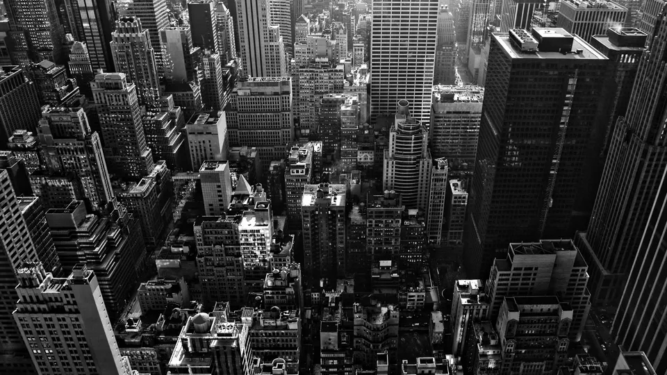 An aerial, black and white view of dense New York City skyscrapers under a bright sky.