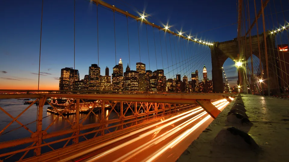 View of the New York City skyline and Brooklyn Bridge at dusk, with light trails from traffic below.