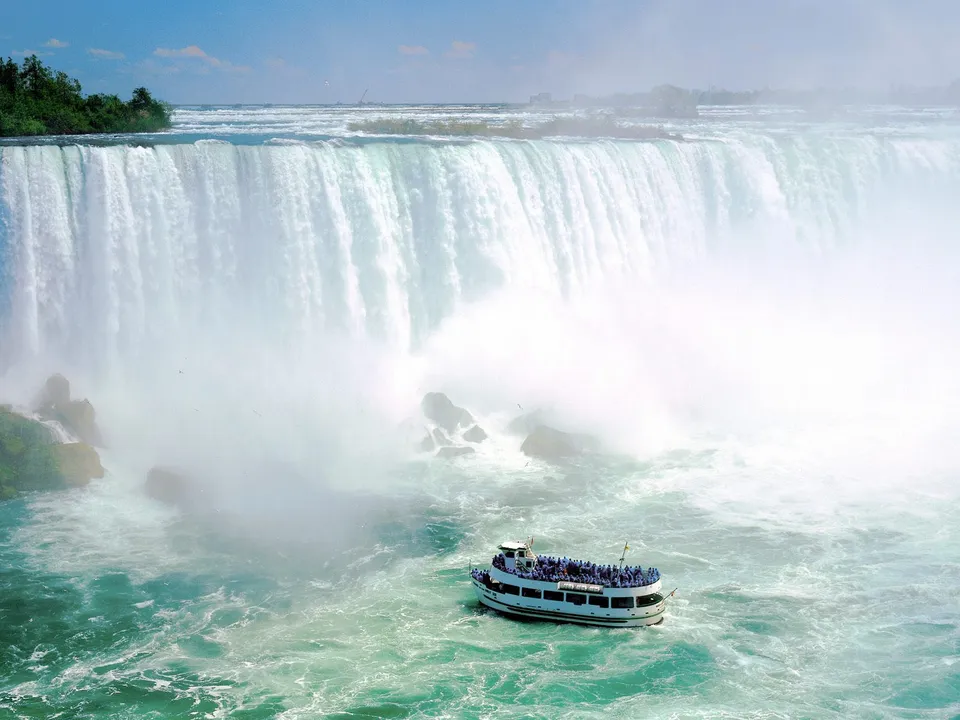 A tour boat filled with people approaches the base of a massive waterfall, surrounded by mist and turbulent water.