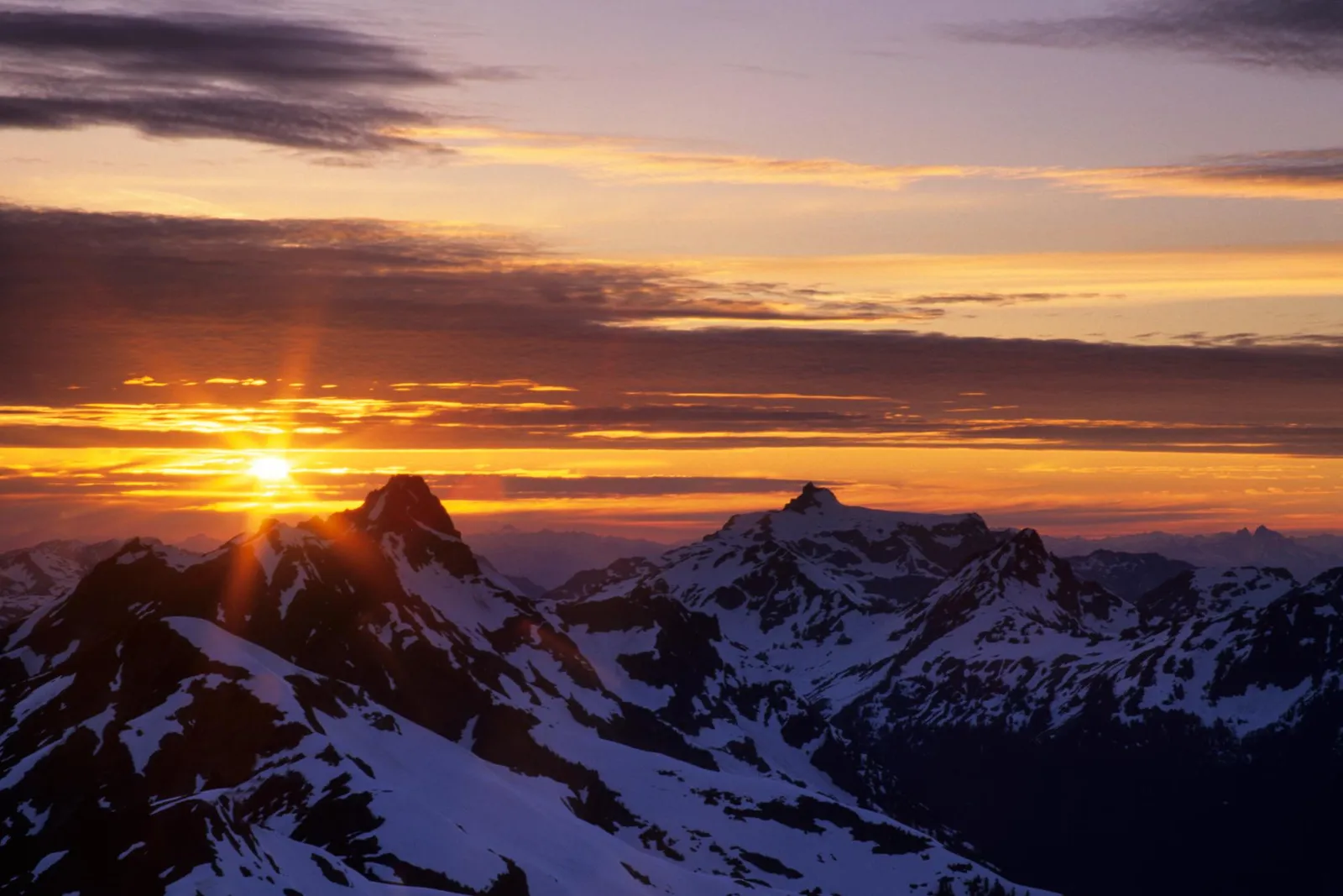 The sun sets behind snow-covered mountains, casting golden light across the sky and peaks.