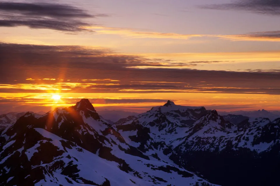 The sun sets behind snow-covered mountains, casting golden light across the sky and peaks.