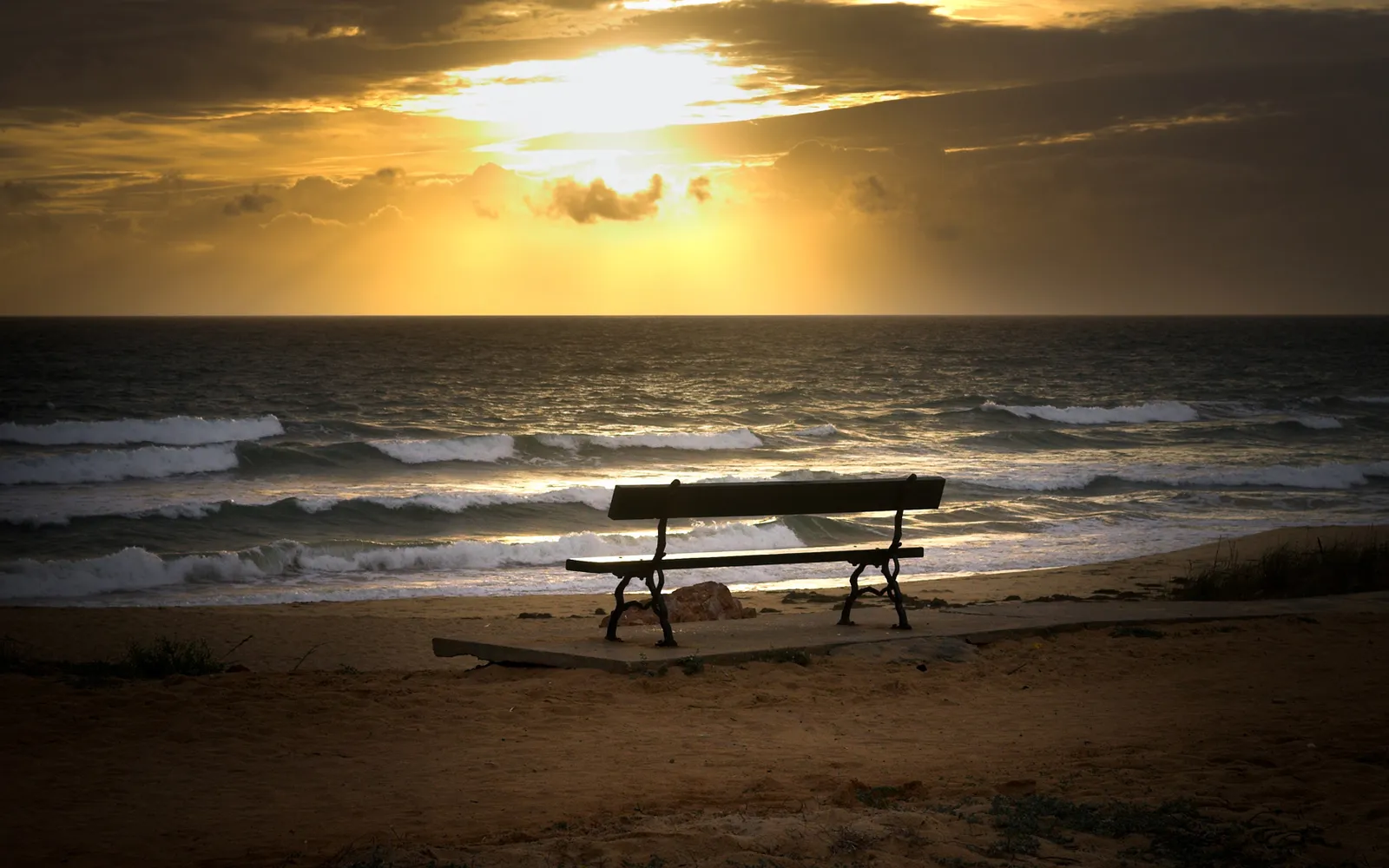 A dark bench sits on a sandy beach facing the ocean as the sun sets.