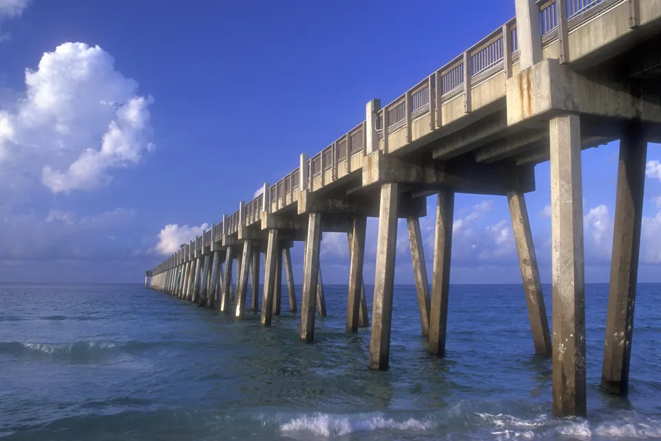 A long concrete pier with wooden railings extends over the blue ocean under a bright blue sky with white clouds.