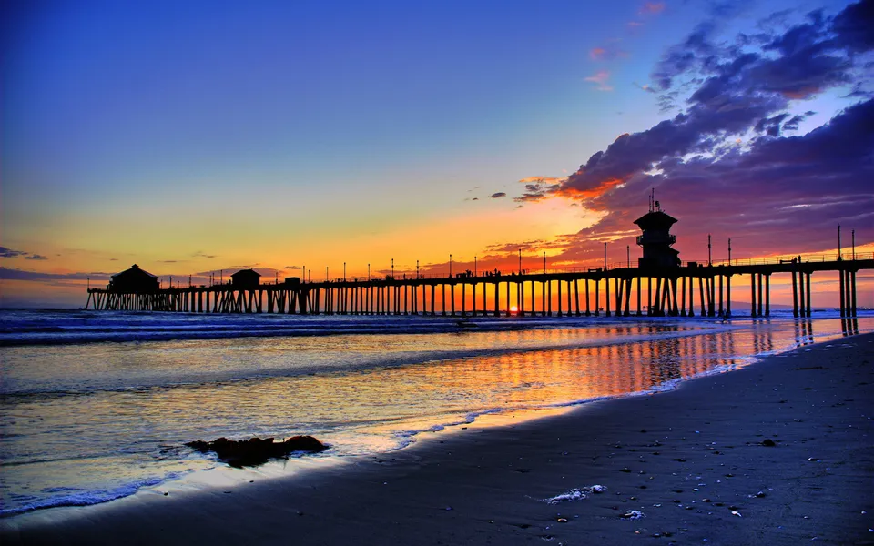 A long pier stretches out over the ocean during a colorful sunset, with waves lapping the shore.
