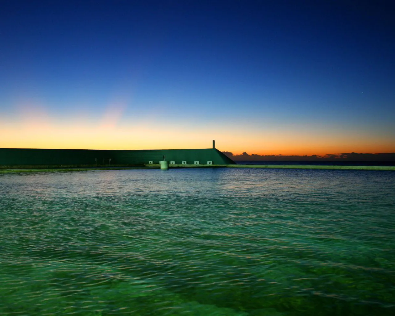 A wide ocean pool with clear green water, bordered by a dark structure, under a gradient sky of blue to orange at dusk.