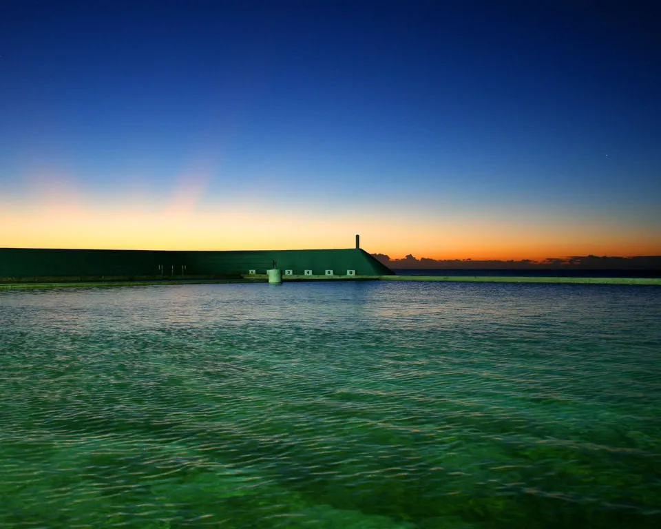 A wide ocean pool with clear green water, bordered by a dark structure, under a gradient sky of blue to orange at dusk.