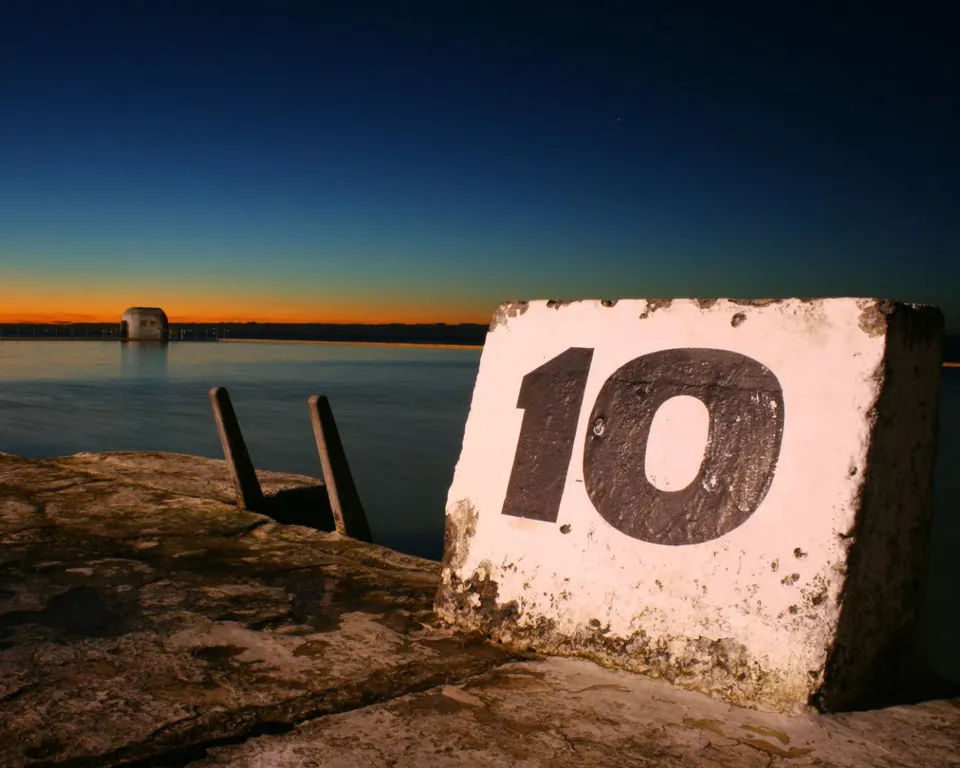 A concrete marker with the number 10 in black stands in the foreground of an ocean pool at dusk.