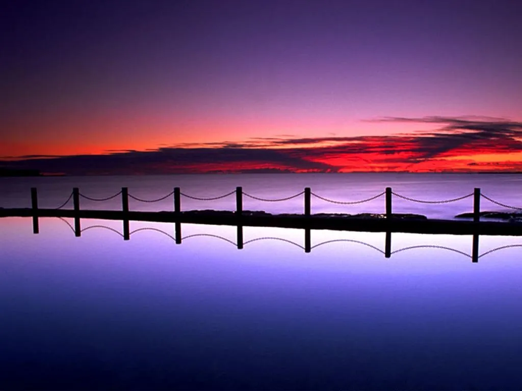 A silhouetted fence in an ocean pool reflects a vibrant sunset sky with red and purple hues.