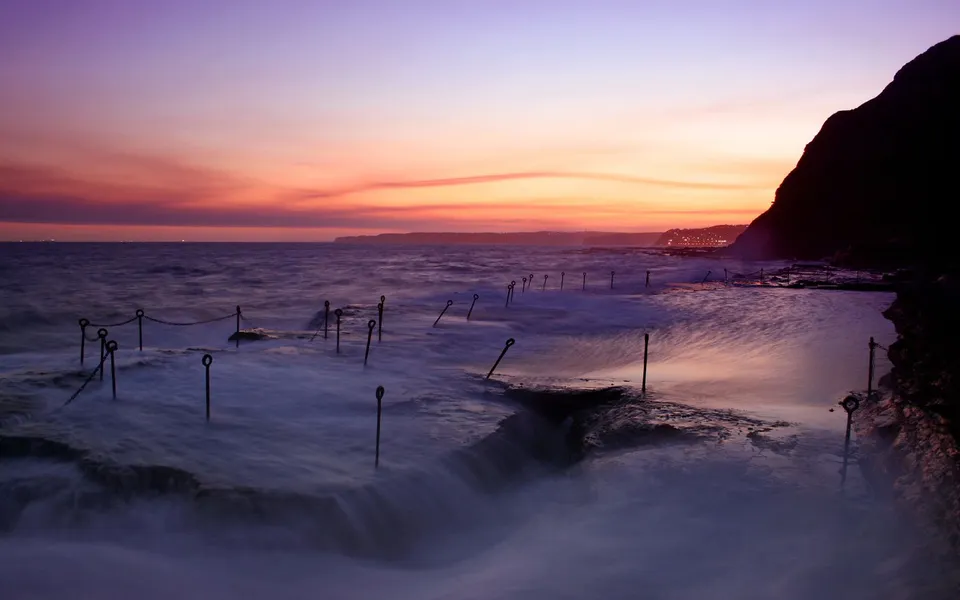 A silken effect of water flows over a rocky ocean pool with metal posts and chains at sunset.