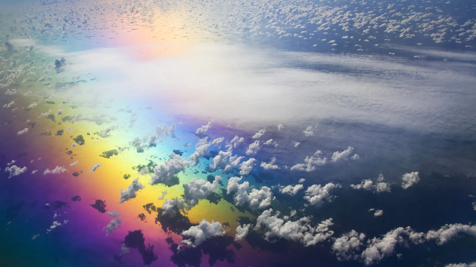 An aerial view of a rainbow reflected on the ocean surface, with scattered clouds above.