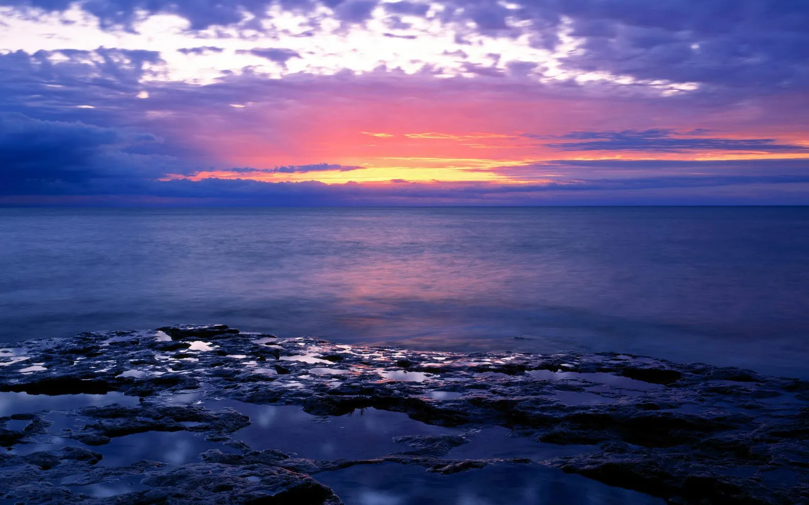 A colorful sunrise paints the sky with pink, orange, and purple hues over a calm ocean, with rocky shore in the foreground.