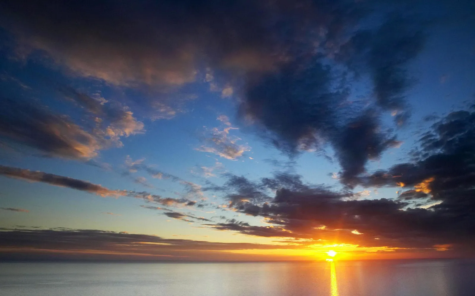 The sun rises over a calm ocean, casting a golden reflection on the water and illuminating colorful clouds.