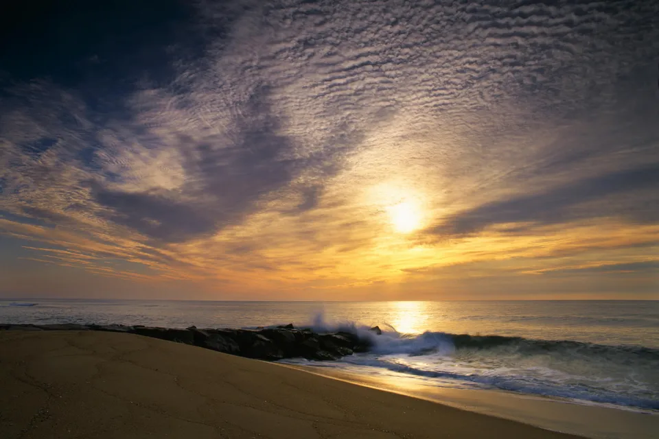 A vibrant sunrise over the ocean, with waves crashing against rocks on a sandy beach.