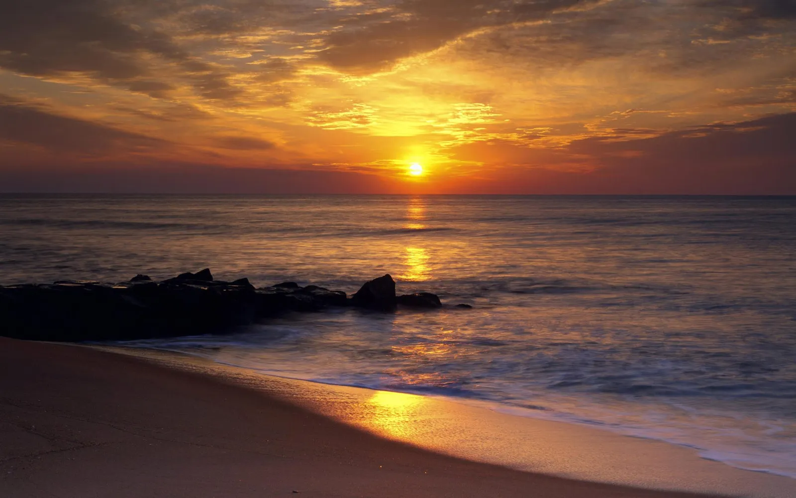A vibrant sunrise over the ocean, with golden light reflecting on the water and a sandy beach in the foreground.