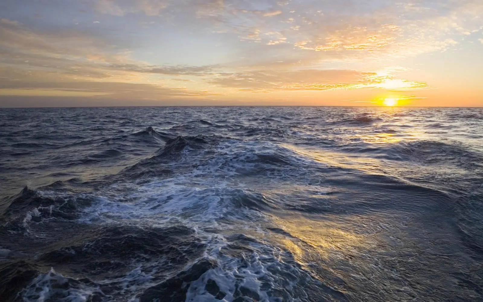 The sun rises over a vast, choppy ocean, casting golden light on the water and clouds.
