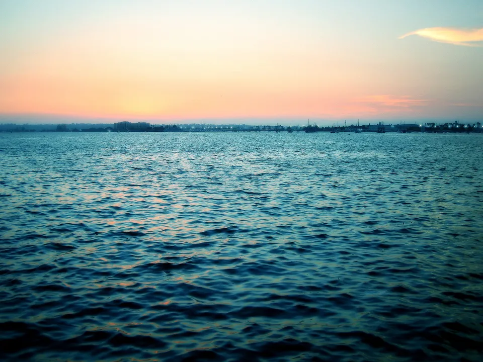A wide expanse of blue water stretches towards a hazy horizon with a soft orange and pink sunset sky.