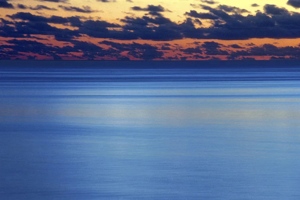 A serene ocean scene at dusk with horizontal lines of water and a band of clouds illuminated by the setting sun.