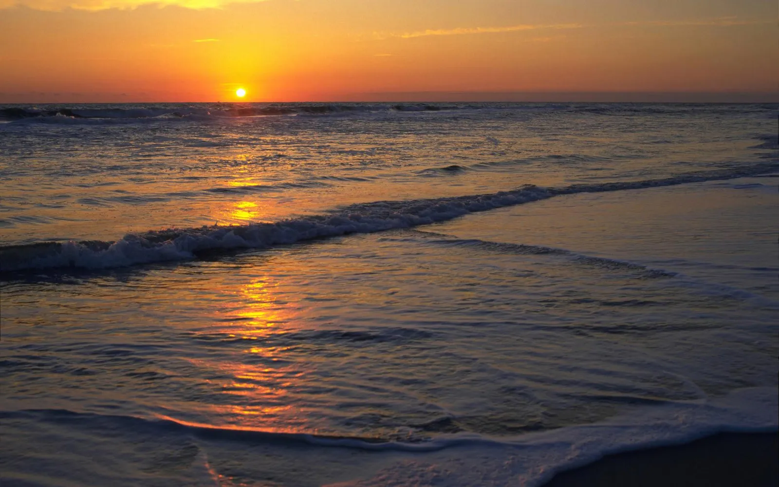 The sun sets over the ocean, casting a golden glow on the water and waves lapping the shore.