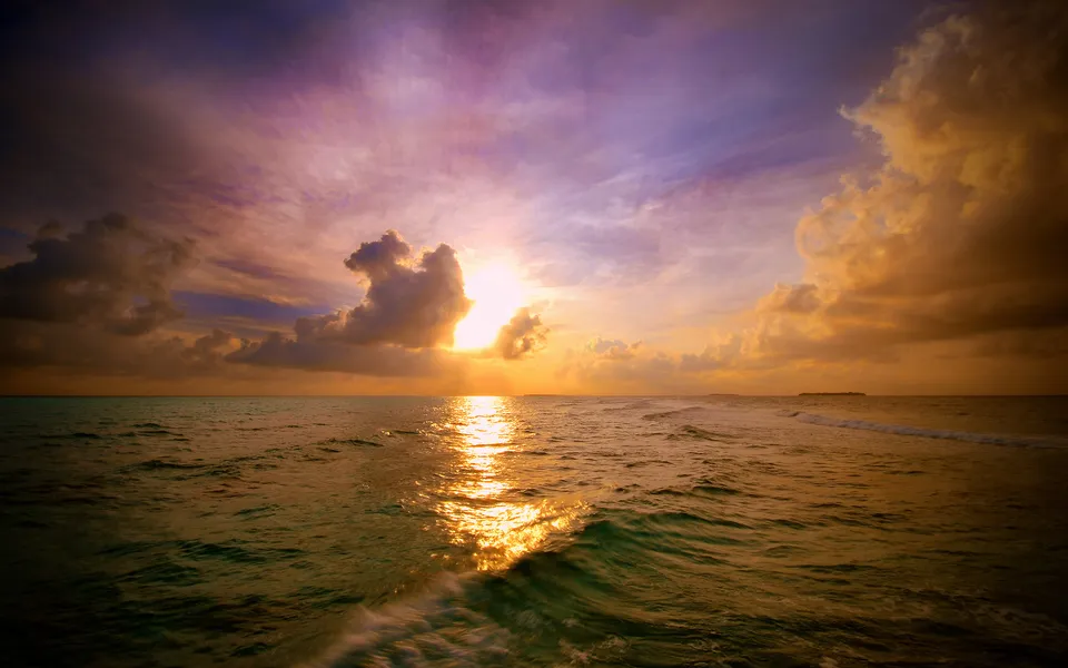 The sun sets over a vast ocean, casting a golden reflection on the rippling water and illuminating dramatic clouds.