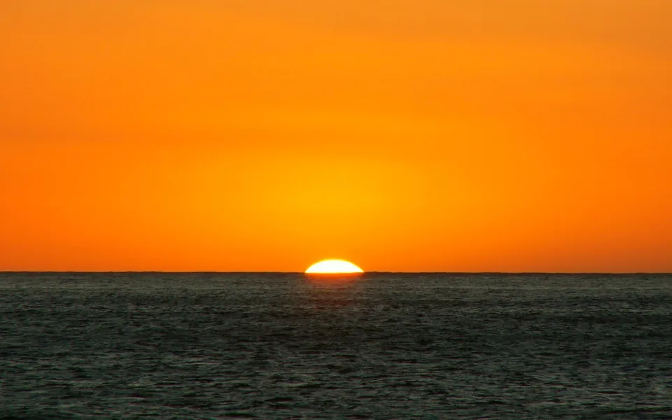 The sun is setting over the ocean, casting a vibrant orange glow across the sky.