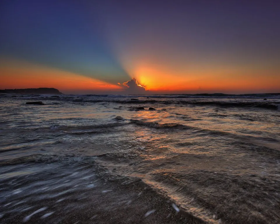 Sun rays burst from behind a cloud during a vibrant sunset over the ocean.