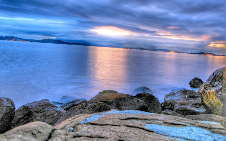 A rocky shoreline meets a calm ocean under a dramatic sunset sky with clouds.