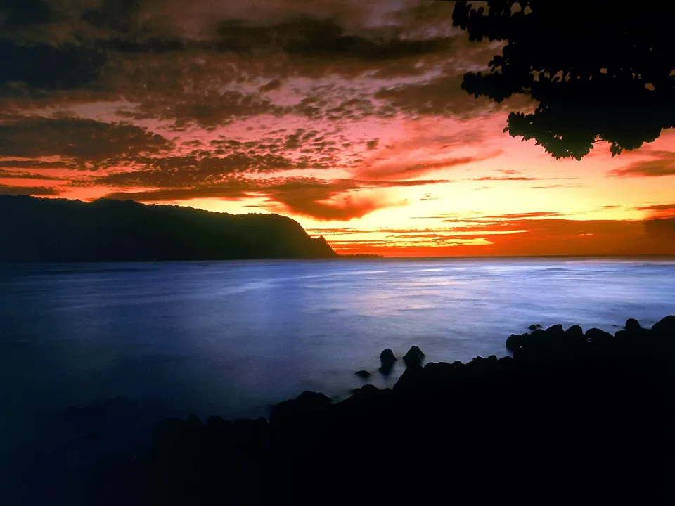 A vibrant sunset paints the sky with orange and pink hues over a calm ocean, with a dark, silhouetted mountain range and rocky shore.
