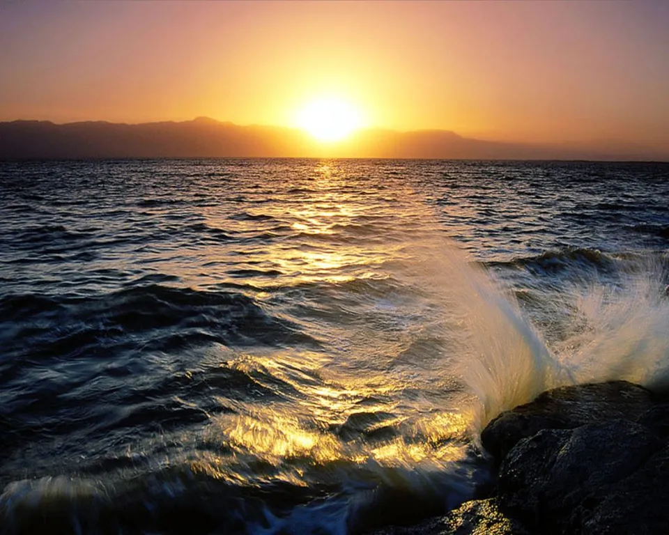 A wave crashes against rocks at sunset, with the sun low on the horizon and casting golden light on the water.