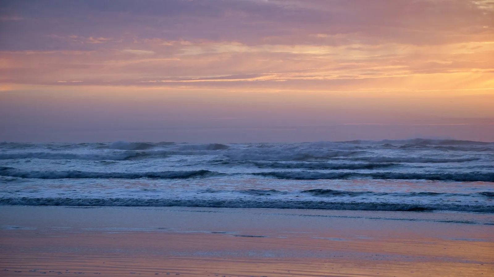 Waves crash on a beach under a pastel-colored sky at sunset.