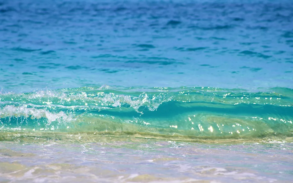 A close-up of a clear turquoise ocean wave breaking gently on a sandy beach.
